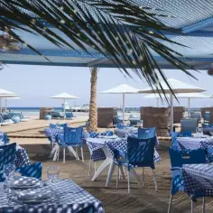 Stoliki z niebiesko-białymi obrusami i krzesłami na plaży w resorcie Pharaoh Azur. W tle leżaki i parasole.