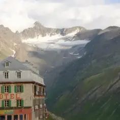 Hotel Belvedere w Szwajcarii, kamienny budynek z zielonymi okiennicami, góruje nad krętą drogą w alpejskim krajobrazie.