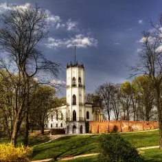 Biały zamek z wieżą na wzgórzu, otoczony drzewami i ceglanym murem. Wiosenna zieleń i żółte krzewy dodają uroku tej opinogórze.
