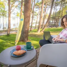 Kobieta pracuje na laptopie przy stoliku z kawą i croissantem. W tle widać morze i domki z wyjściem na plażę.