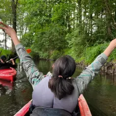 Rzeka Reda w otoczeniu zieleni. Dwie osoby płyną kajakami, jedna z nich radośnie unosi ręce w geście zwycięstwa.