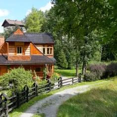 Drewniany dom z charakterystyczną architekturą, otoczony zielenią. Widać fragmenty innych budynków, sugerujące spokojne zakopane dzielnice.