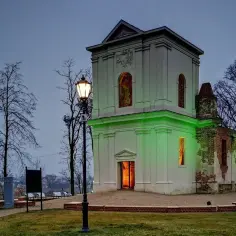 Zabytkowa kaplica w Piaskach Lubelskich, częściowo odrestaurowana, oświetlona zielonym światłem. Wokół drzewa i latarnie.