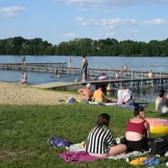 Ludzie odpoczywają nad jeziorem, niektórzy kąpią się, inni opalają na plaży i trawie. W tle widać drewniane molo. To idealne miejsce na wypoczynek, jak kamień krajeński.