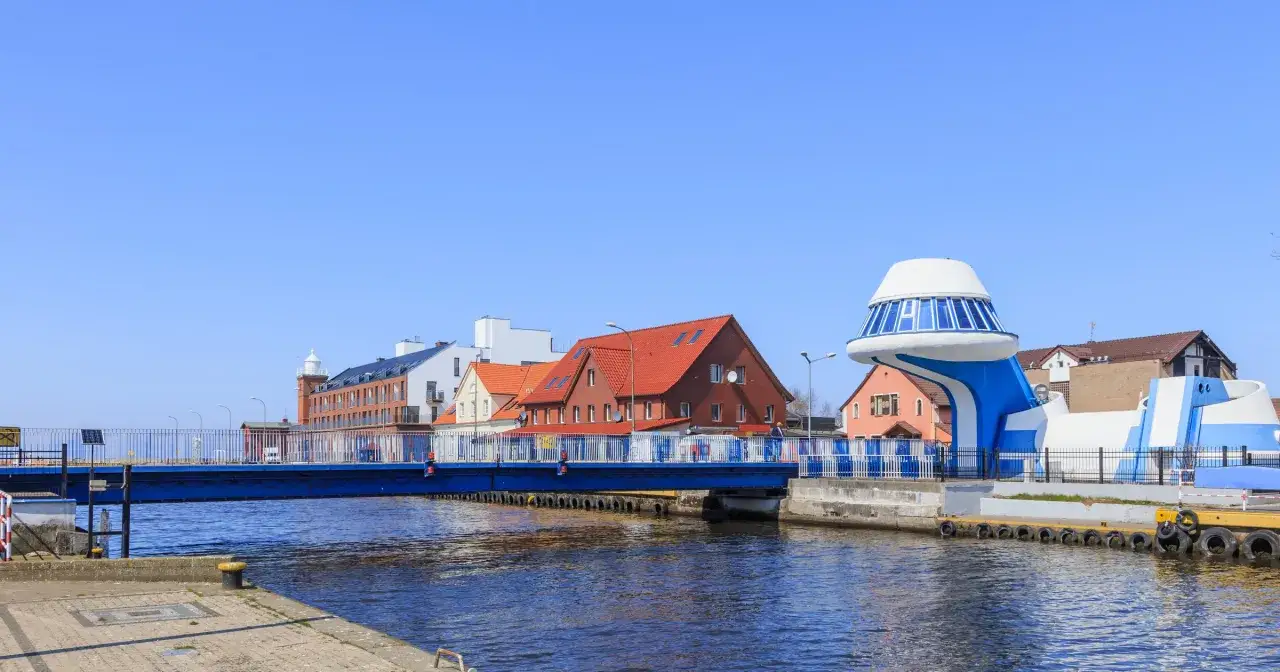 Niebieski most nad kanałem, budynki z czerwonymi dachami i futurystyczna budowla tworzą letni sen nad wodą.