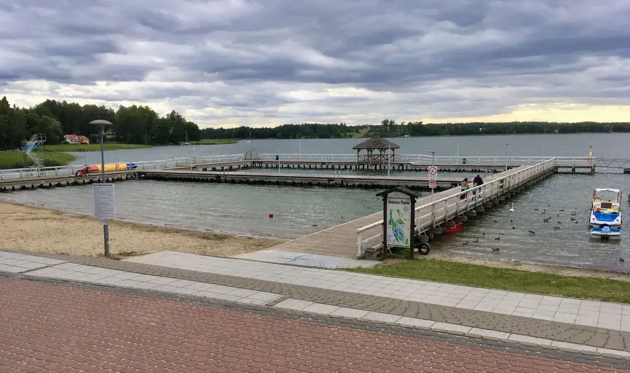 Miasto nad jeziorem Skiertąg z drewnianymi pomostami, plażą i łódkami. Kaczki pływają w wodzie.