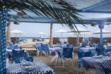 Stoliki z niebiesko-białymi obrusami i krzesłami na plaży w resorcie Pharaoh Azur. W tle leżaki i parasole.