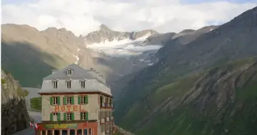 Hotel Belvedere w Szwajcarii, kamienny budynek z zielonymi okiennicami, góruje nad krętą drogą w alpejskim krajobrazie.