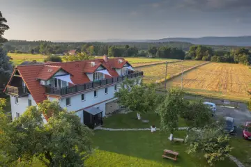 Malowniczy dom z czerwonym dachem i balkonami, otoczony zielonym ogrodem i polami. Idealne miejsce na wypoczynek w spokojnej okolicy.