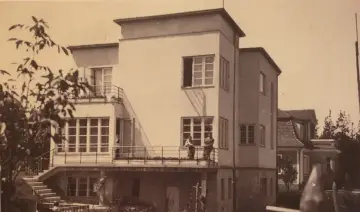 Dwaj mężczyźni stoją na tarasie modernistycznej willi w Grabówku. W tle widać inne budynki i drzewa.