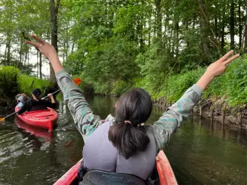 Rzeka Reda w otoczeniu zieleni. Dwie osoby płyną kajakami, jedna z nich radośnie unosi ręce w geście zwycięstwa.