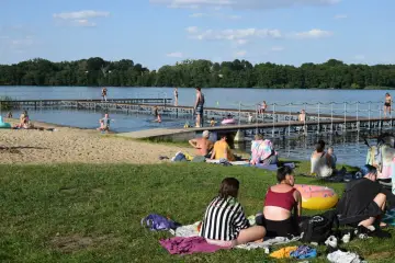 Ludzie odpoczywają nad jeziorem, niektórzy kąpią się, inni opalają na plaży i trawie. W tle widać drewniane molo. To idealne miejsce na wypoczynek, jak kamień krajeński.