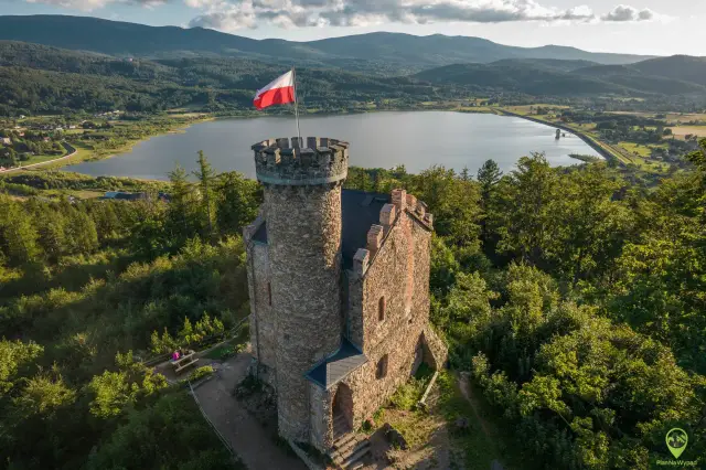 Kamienna wieża z polską flagą na tle jeziora i gór. To widok z Sudetów, gdzie można podziwiać piękne krajobrazy.