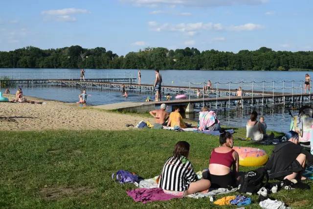 Ludzie odpoczywają nad jeziorem, niektórzy kąpią się, inni opalają na plaży i trawie. W tle widać drewniane molo. To idealne miejsce na wypoczynek, jak kamień krajeński.